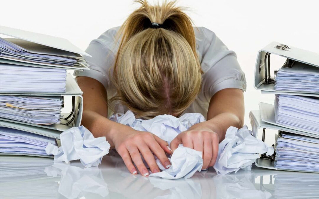 a woman experiencing burnout buries her head in a pile of crumpled papers between two large stacks of binders.
