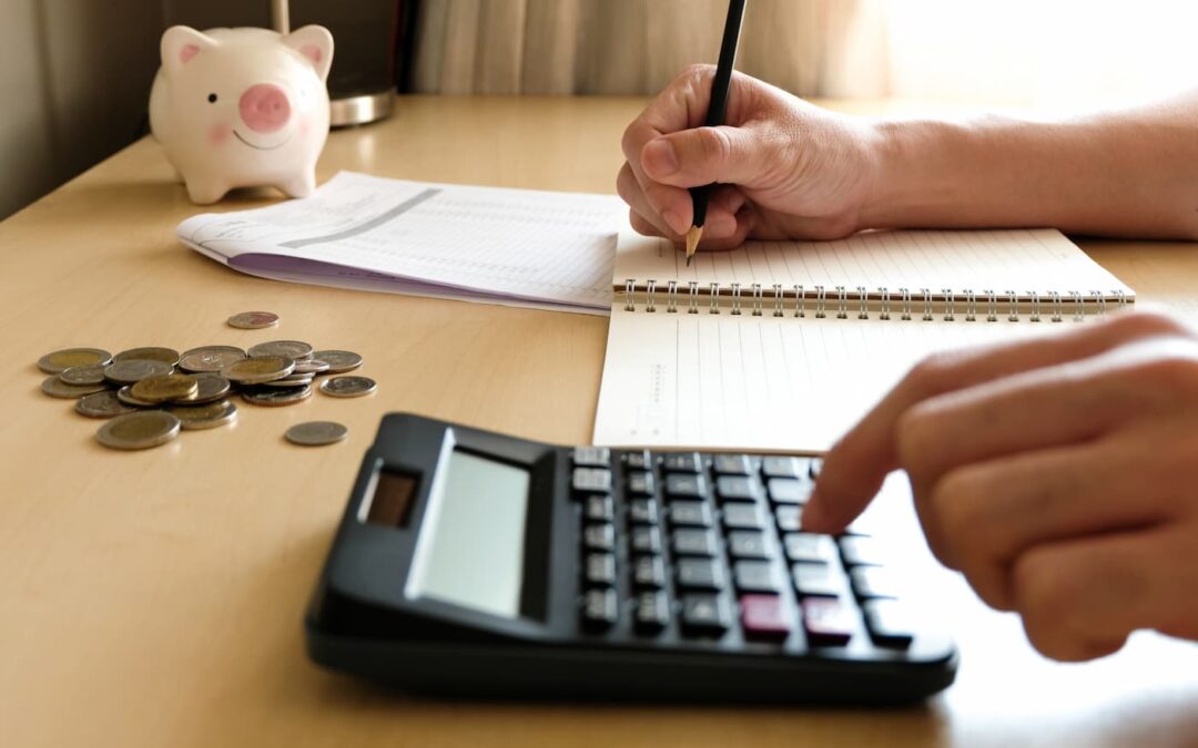 a person sitting at a desk using a pen, pencil, and calculator to add up their property management costs. A piggy bank and coins also sit on the desk.