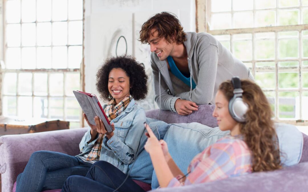 Two women sit on a purple couch in a room filled with natural light. A man, who is their guest, looks over one's of the woman's shoulder to read something off her tablet.