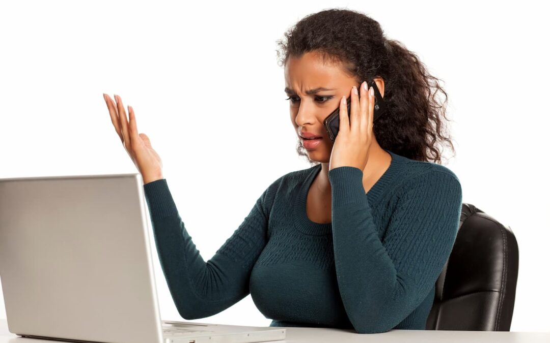 A women wearing a black sweater sitting in front a loptop computer makes a complaint into her cell phone.